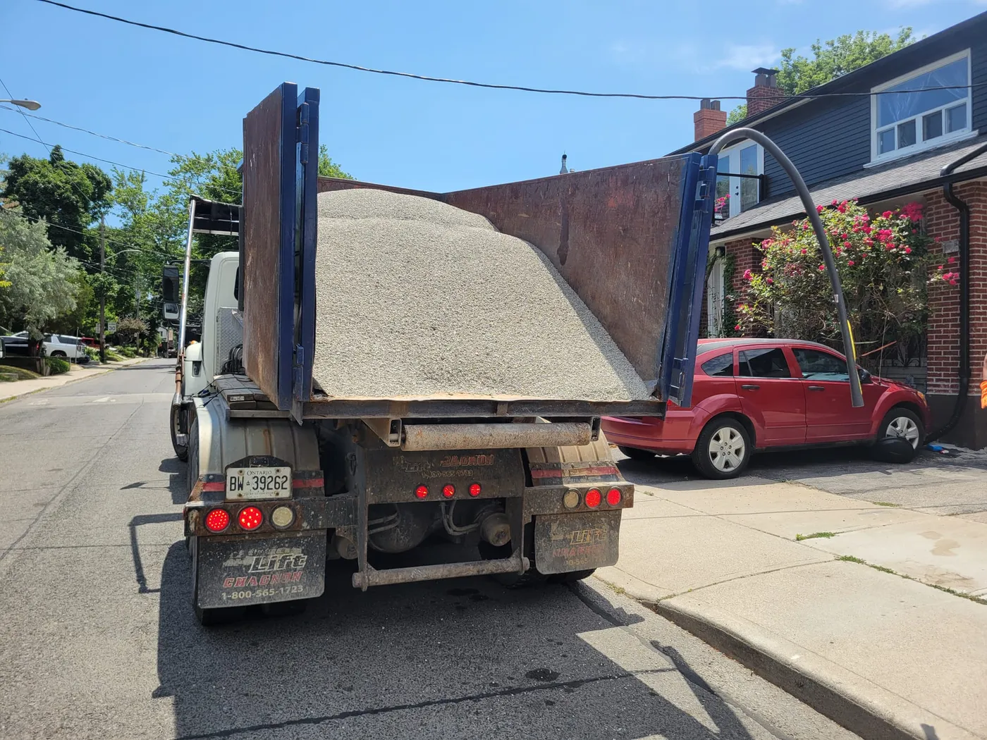 Roll-off truck carrying light-colored aggregate, parked on a residential street beside a brick house