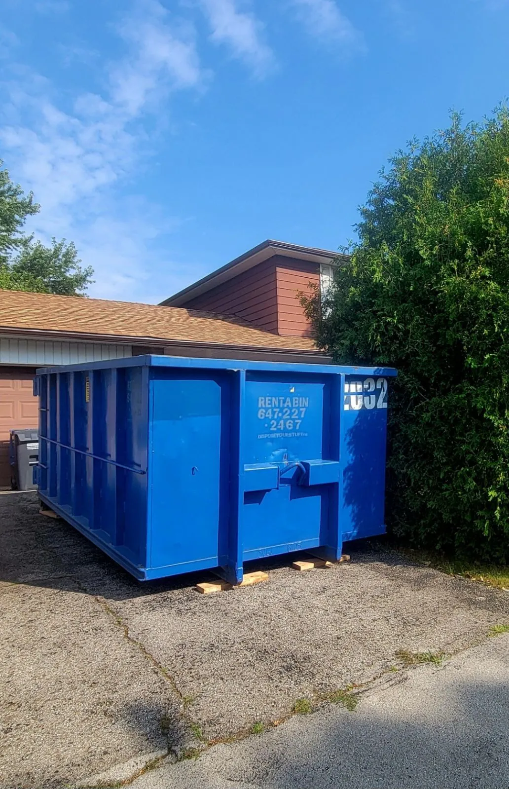 A blue roll-off bin on a residential driveway protected by wooden planks.