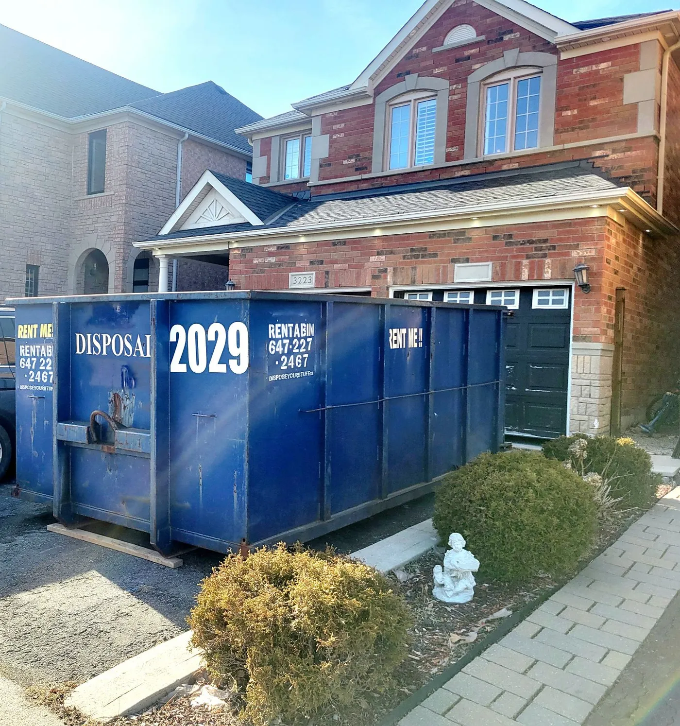Blue Dispose Your Stuff roll-off bin with company branding and phone number parked on a brick home's driveway